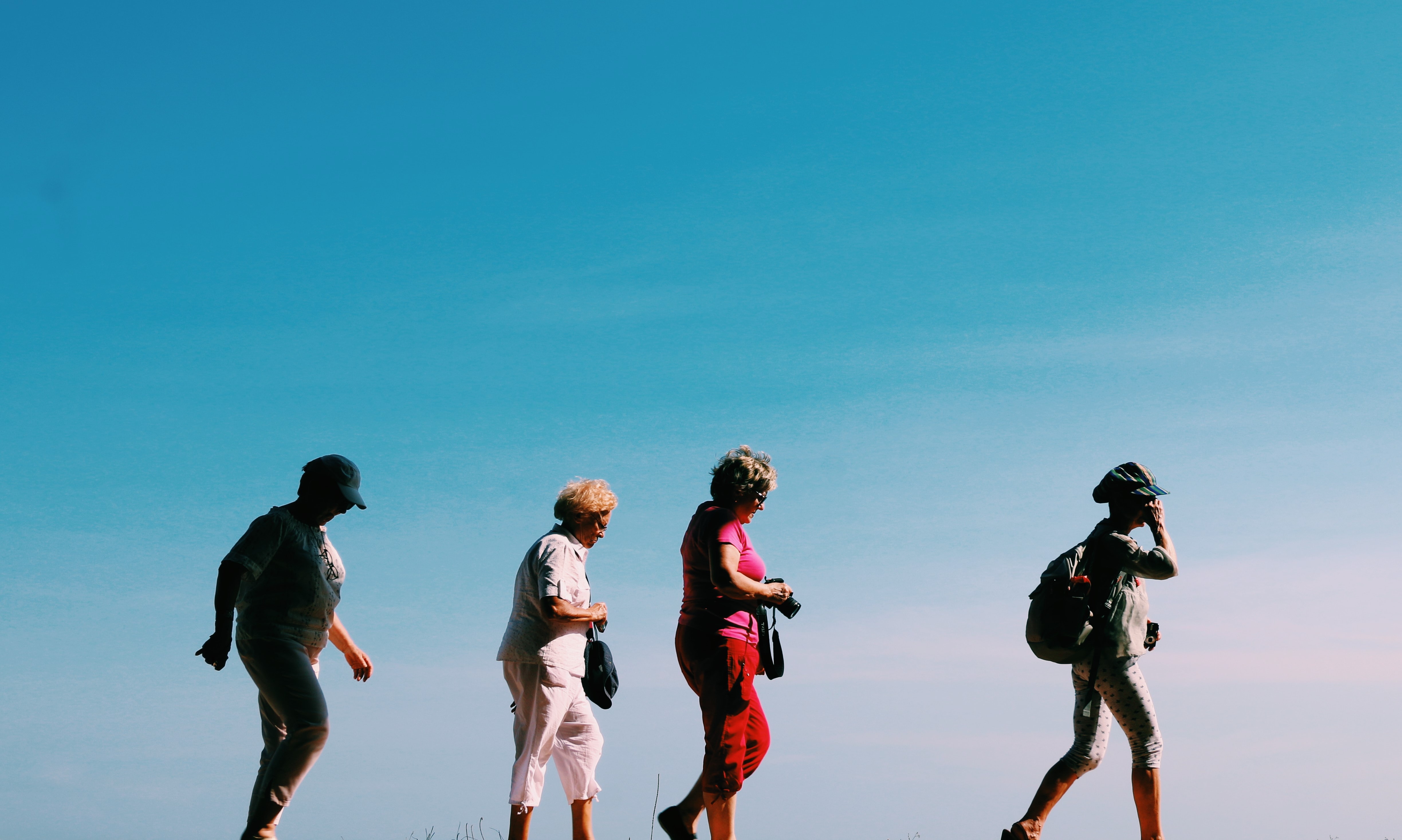 Fyra äldre personer med telefoner och kameror går på rad med blå himmel som bakgrund