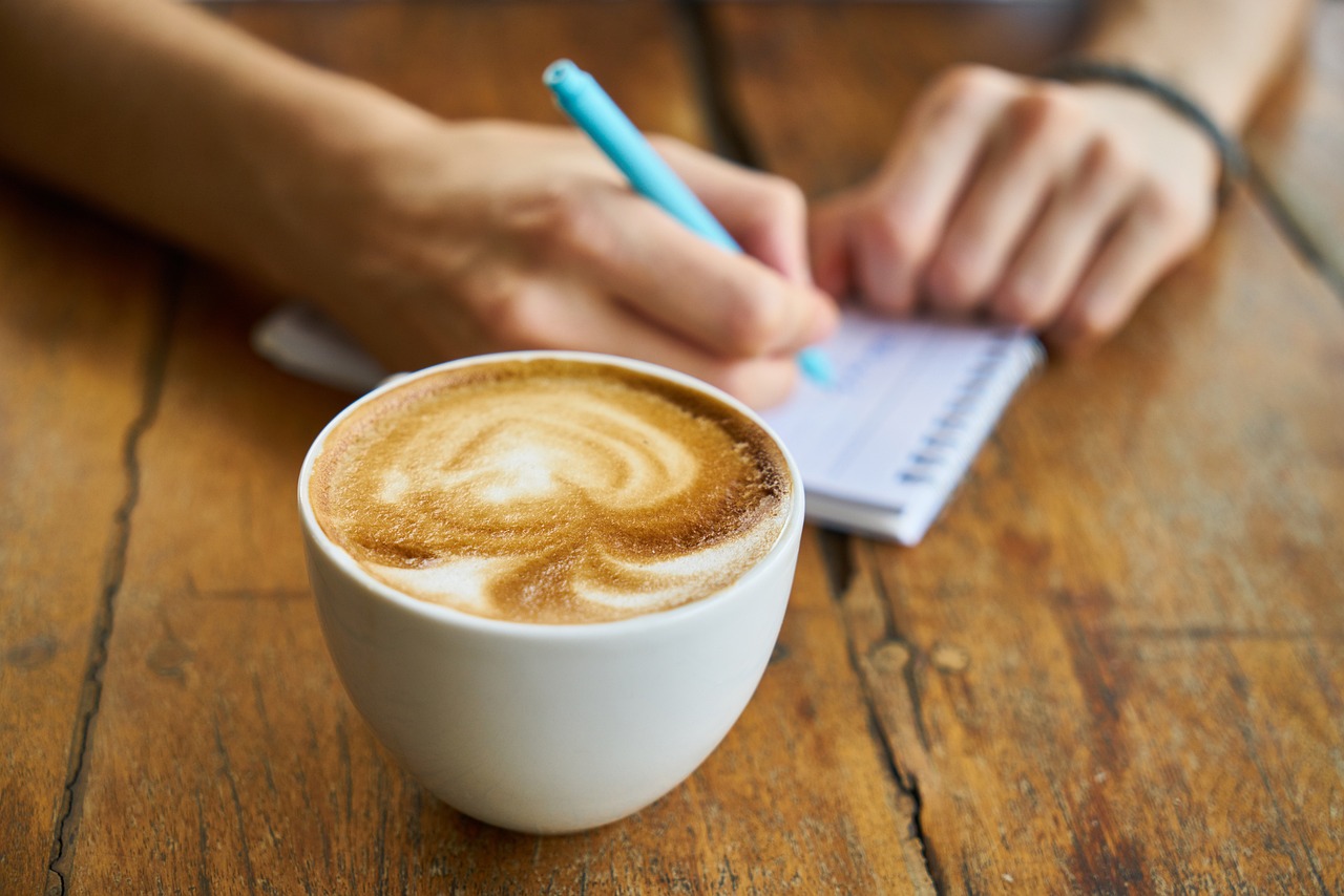 En kaffekopp och händer som håller i en penna och ett kollegieblock. 