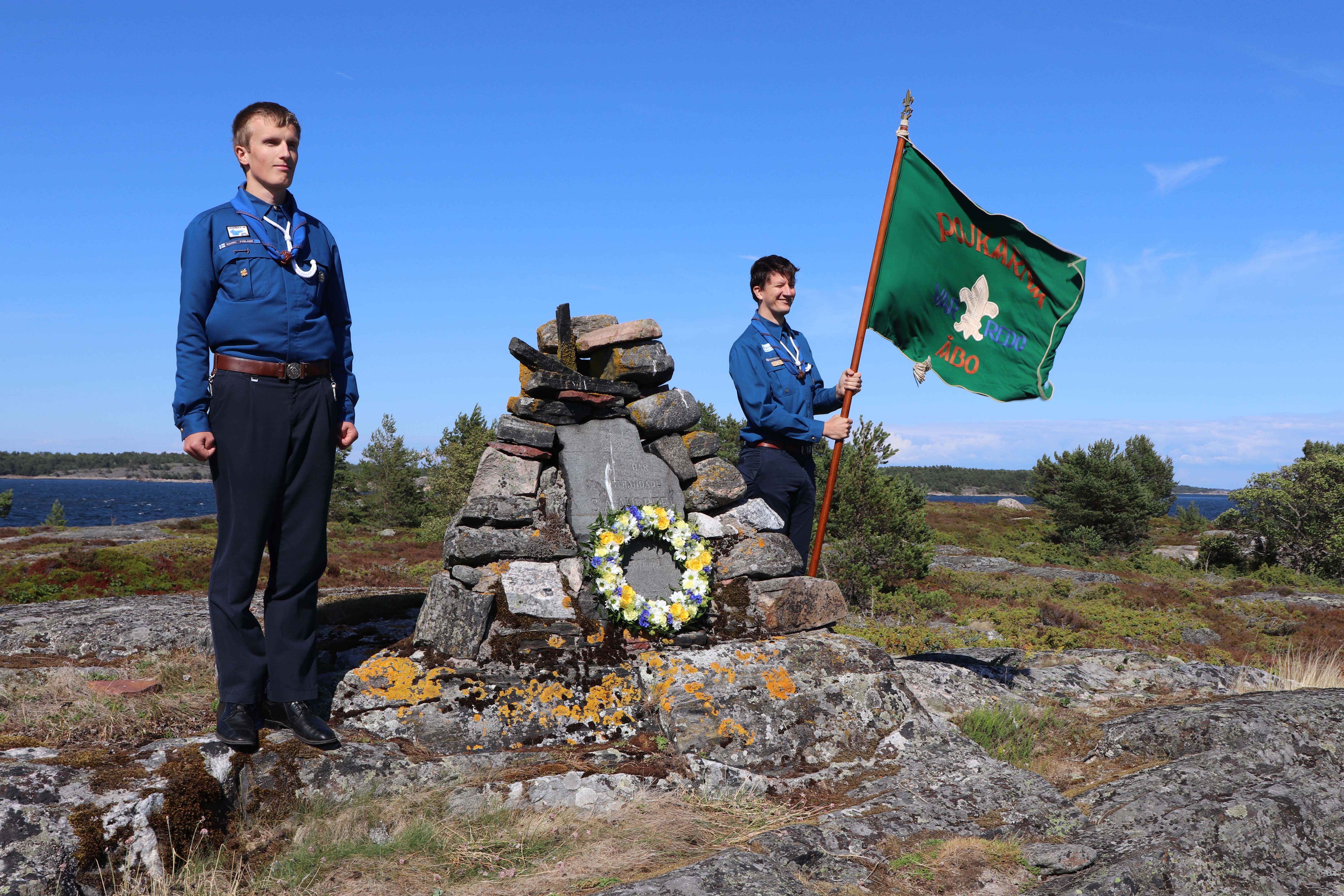 Två personer står vid ett minnesmärke och håller en flagga